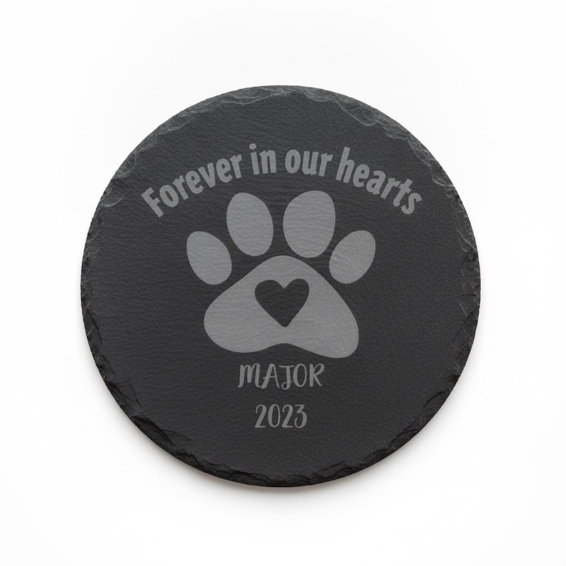 May include: A round black slate coaster with a rough, natural edge. The surface is engraved with a paw print containing a small heart, the text "Forever in our hearts", the name "MAJOR", and the year "2023" in a light gray color.