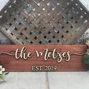 May include: A wooden sign with the text "the Metzes EST. 2019" in gold lettering. The sign is a dark reddish-brown color.