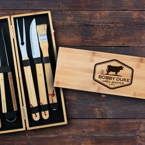 May include: A wooden box set containing a grill spatula, tongs, fork, knife, and a basting brush. The box is engraved with the text "Bobby Duke Grill Master" and a silhouette of a cow.
