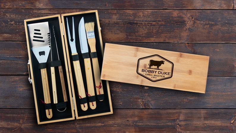 May include: A wooden box set containing a grill spatula, tongs, fork, knife, and a basting brush. The box is engraved with the text "Bobby Duke Grill Master" and a silhouette of a cow.