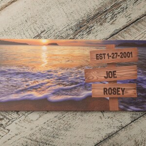 May include: A wooden sign with a sunset beach background. The sign has three wooden planks with the text "EST 1-27-2001", "JOE", and "ROSE" on them.