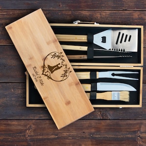 May include: A bamboo barbecue tool set in a wooden case. The case lid is engraved with a couple silhouette inside a heart-shaped floral design and the names "Rachel & Mike" with the year "est. 2018". The set includes a spatula, fork, knife, brush, and tongs.