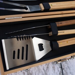 May include: A wooden carrying case with a black interior holds a set of grilling tools. The tools include a spatula with a bottle opener, a pair of tongs, a grill fork, and a basting brush.