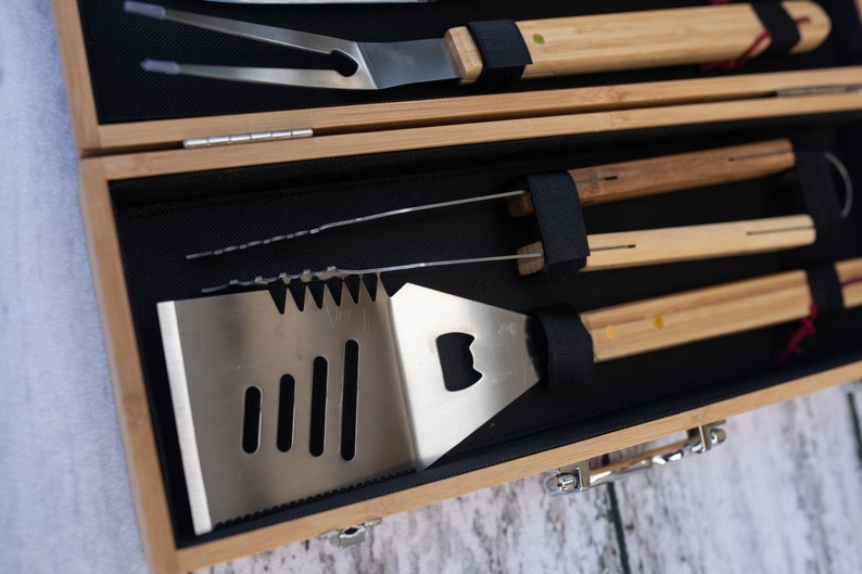 May include: A wooden carrying case with a black interior holds a set of grilling tools. The tools include a spatula with a bottle opener, a pair of tongs, a grill fork, and a basting brush.
