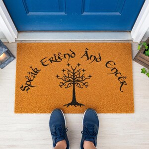 A brown doormat with the text "Speak En'er" and a tree design, placed on a concrete floor next to a window.