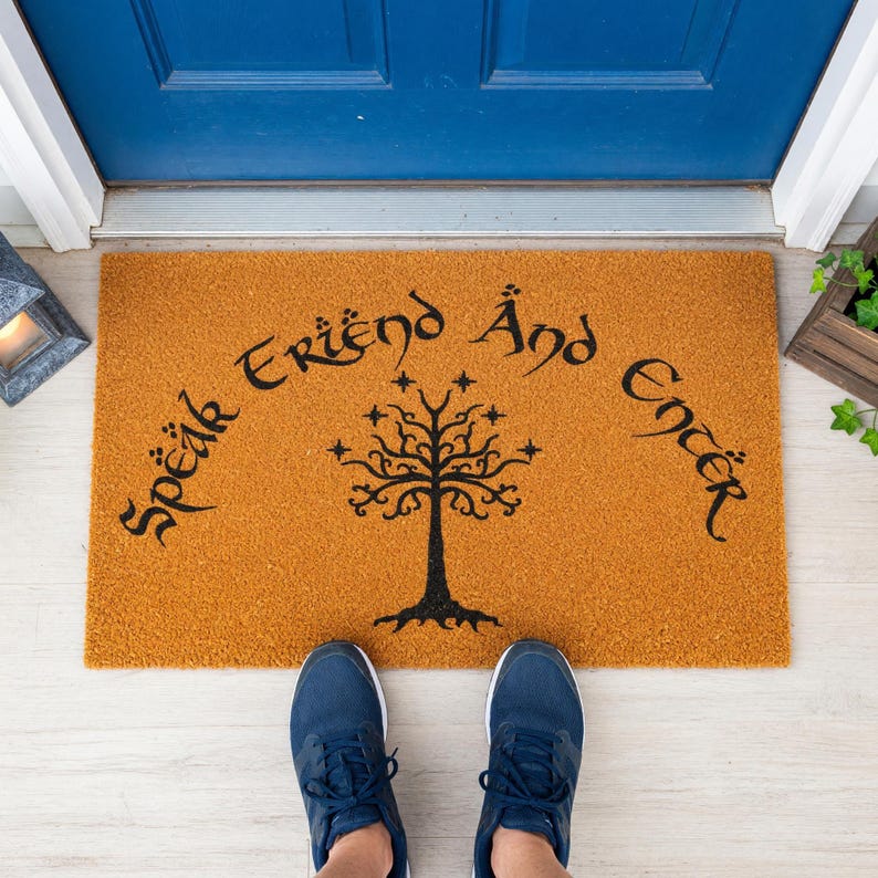 A brown doormat with the text "Speak En'er" and a tree design, placed on a concrete floor next to a window.