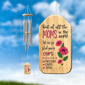 May include: A wooden sign with the text "Out of all the MOMS in the World We are so glad you're ours" with pink flowers and the names Matilda, Carol, and John. A silver wind chime hangs above the sign.