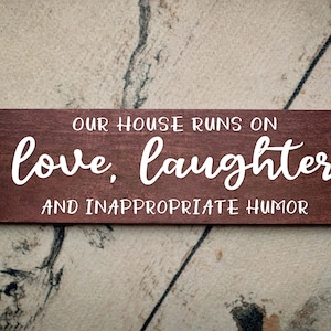 May include: A brown wooden sign with white text that reads "Our house runs on love, laughter and inappropriate humor."