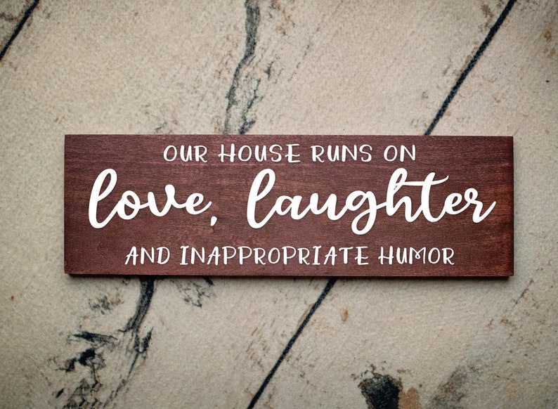 May include: A brown wooden sign with white text that reads "Our house runs on love, laughter and inappropriate humor."