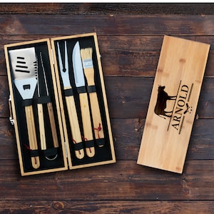 May include: A wooden box set with a personalized engraved cutting board and a set of five grilling tools. The tools include a spatula, tongs, a fork, a knife, and a basting brush. The cutting board has a cow silhouette and the name "ARNOLD" engraved on it.