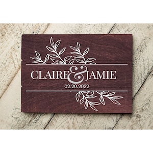May include: A wooden sign with a burgundy stain, featuring a white floral design and the text "CLAIRE & JAMIE 02.20.2022".