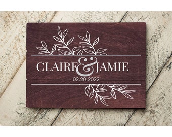 May include: A wooden sign with a burgundy stain, featuring a white floral design and the text "CLAIRE & JAMIE 02.20.2022".