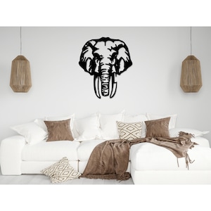 May include: Black metal wall decor featuring a silhouette of an elephant's head with large tusks. The decor is mounted on a white wall above a white couch with brown throw pillows and a brown blanket.