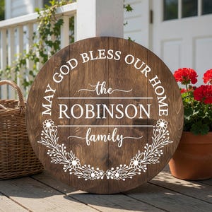 May include: A round dark brown wooden sign featuring white text that reads MAY GOD BLESS OUR HOME, the ROBINSON family, and a decorative floral wreath design. The sign has a visible wood grain texture and is displayed on a wooden porch floor.