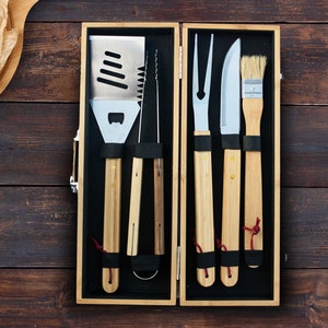 May include: A set of six grilling tools with wooden handles, including a spatula, tongs, a basting brush, a fork, and a knife, all neatly arranged in a wooden box.