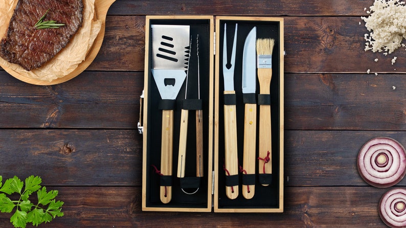 May include: A set of six grilling tools with wooden handles, including a spatula, tongs, a basting brush, a fork, and a knife, all neatly arranged in a wooden box.