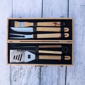 May include: A set of grilling tools with wooden handles in a wooden carrying case. The tools include a spatula, tongs, a fork, a knife, and a brush.