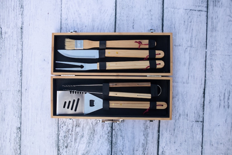 May include: A set of grilling tools with wooden handles in a wooden carrying case. The tools include a spatula, tongs, a fork, a knife, and a brush.