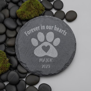 May include: A round, dark gray slate stone engraved with the text "Forever in our hearts" above a paw print graphic containing a small heart. Below the paw print, the name "MAJOR" is engraved, followed by the year "2023" in a simple font.