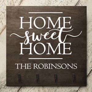 May include: A brown wooden key holder with the text "Home sweet home" and "The Robinsons" in white lettering. The key holder has five hooks for hanging keys.