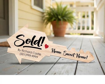 Może przedstawiać: Jasny drewniany znak w kształcie dużego klucza do domu. Napis brzmi Sold! By Richard Castle (501)999-9999 oraz Home Sweet Home! z małą czerwoną ikoną serca. Znak ma naturalne usłojenie drewna i czarne litery.