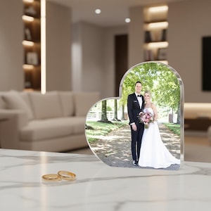 May include: A heart-shaped photo frame with a picture of a couple in wedding attire. The frame is clear and stands on a white surface. Two gold wedding rings are in the foreground.