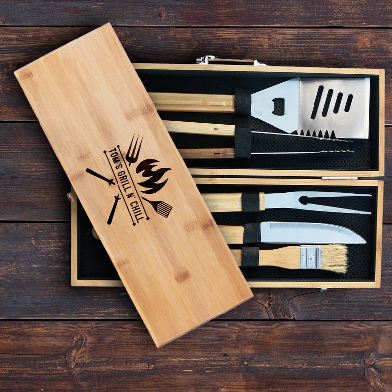 May include: A bamboo grilling set with a wooden carrying case. The case has a personalized inscription that reads "Tom's Grill N Chill". The set includes a spatula, tongs, a grill brush, a fork, and a knife.