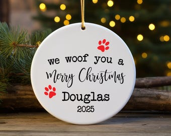 Peut inclure: Ornement de Noël rond et blanc avec l'inscription "we woof you a Merry Christmas" en noir. Il présente deux empreintes de pattes rouges et le nom "Douglas". L'année "2025" est imprimée en dessous. Une ficelle dorée est attachée pour l'accrocher.