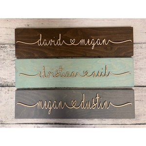 Puede incluir: Tres letreros de madera con los nombres de parejas y un s&iacute;mbolo de coraz&oacute;n entre ellos. El letrero superior es marr&oacute;n con los nombres "david & megan". El letrero del medio es azul claro con los nombres "christian & neil". El letrero inferior es gris con los nombres "megan & dustin".