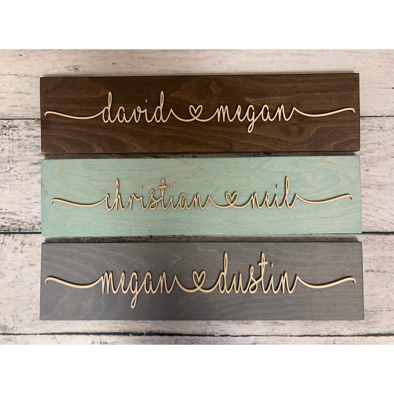 Puede incluir: Tres letreros de madera con los nombres de parejas y un s&iacute;mbolo de coraz&oacute;n entre ellos. El letrero superior es marr&oacute;n con los nombres "david & megan". El letrero del medio es azul claro con los nombres "christian & neil". El letrero inferior es gris con los nombres "megan & dustin".