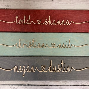 Puede incluir: Tres letreros de madera con los nombres de parejas escritos en cursiva con un s&iacute;mbolo de coraz&oacute;n entre los nombres. El letrero superior es rojo, el letrero del medio es azul claro y el letrero inferior es gris.