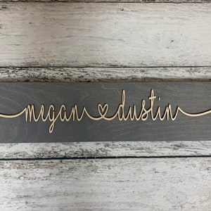 Puede incluir: Un letrero de madera gris con los nombres "megan" y "dustin" escritos en cursiva con un s&iacute;mbolo de coraz&oacute;n entre ellos.
