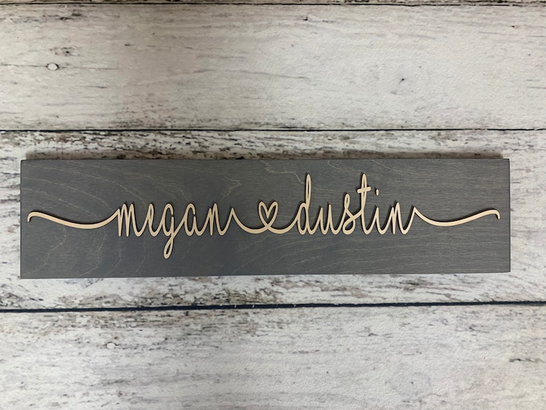 Puede incluir: Un letrero de madera gris con los nombres "megan" y "dustin" escritos en cursiva con un s&iacute;mbolo de coraz&oacute;n entre ellos.