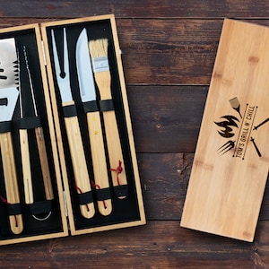 May include: A bamboo grilling tool set with a wooden carrying case. The set includes a spatula, tongs, a fork, a knife, and a basting brush. The case is engraved with the text "Tom's Grill N Chill" and a design of crossed grilling tools and a flame.