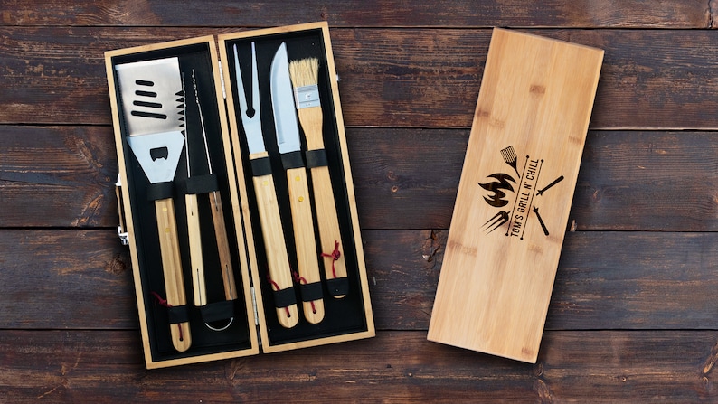May include: A bamboo grilling tool set with a wooden carrying case. The set includes a spatula, tongs, a fork, a knife, and a basting brush. The case is engraved with the text "Tom's Grill N Chill" and a design of crossed grilling tools and a flame.