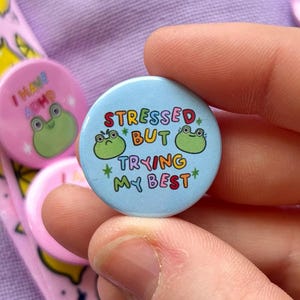 May include: A selection of colourful pin badges with diverse designs and text. One badge says "Stressed but trying my best" with frog illustrations. Other badges feature a strawberry, a flower, and a heart. A pink lanyard with lemon graphics is also visible.