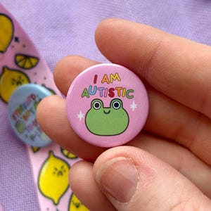 Könnte beinhalten: Ein rosa Knopf mit einem grünen Frosch und dem Text "I AM AUTISTIC" in Regenbogenfarben. Der Knopf wird von einer Hand gehalten.