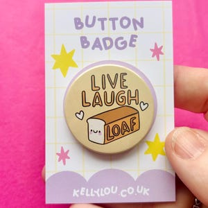 May include: A round button badge on a white display card with a grid pattern, yellow stars, and purple clouds. The badge features a tan background with the text "LIVE LAUGH LOAF" and a cartoon bread loaf with a face. The card text reads "BUTTON BADGE" and "KELLYLOU.CO.UK".