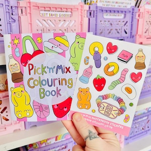 May include: A person holds a "Pick'n Mix Colouring Book" and a sheet of stickers with food-themed designs. The book features a bear, a cherry, and other graphics. The background shows pastel pink and purple storage crates and other books.