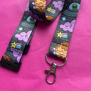 May include: A lanyard featuring a repeating pattern of cartoon planets, clouds, stars, and UFOs on a dark grey background. It has a black plastic clip and a silver metal clasp. The design has a cute, whimsical space theme.