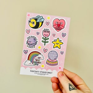 May include: A pink sticker sheet with a variety of colourful stickers featuring positive messages and illustrations. The stickers include a bee, a flower, a rainbow, a star, a heart, and a planet. The text on the stickers includes "Bee Kind", "Bloom", "Give Yourself Space", "You're A Star", "Love Yourself", "You Deserve Joy", and "Make Your Future Better Than Your Past".