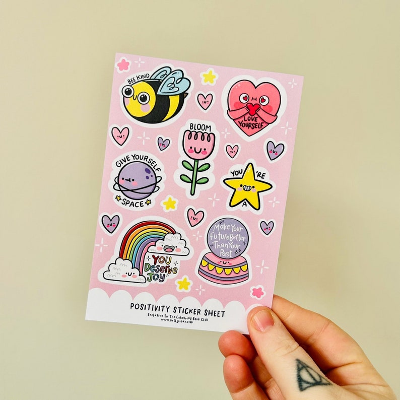May include: A pink sticker sheet with a variety of colourful stickers featuring positive messages and illustrations. The stickers include a bee, a flower, a rainbow, a star, a heart, and a planet. The text on the stickers includes "Bee Kind", "Bloom", "Give Yourself Space", "You're A Star", "Love Yourself", "You Deserve Joy", and "Make Your Future Better Than Your Past".