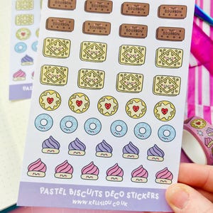 Könnte beinhalten: Ein Bogen mit dekorativen Aufklebern im Pastell-Keks-Design. Die Aufkleber zeigen verschiedene Keksdesigns, darunter Bourbon-Kekse, herzförmige Kekse und Cupcakes. Der Text "PASTEL BISCUITS DECO STICKERS" und die Website "WWW.KELLYLOU.CO.UK" sind sichtbar.