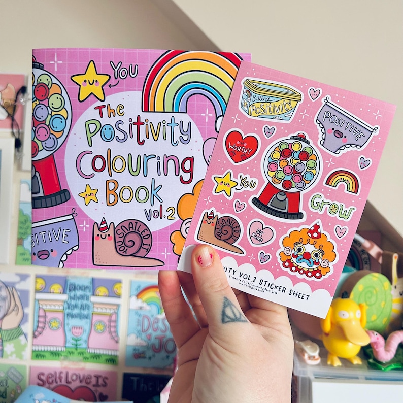 May include: A pink and white colouring book with a rainbow and a gumball machine on the cover. The title is "The Positivity Colouring Book Vol. 2". A sticker sheet with a pink background and various colourful stickers is also shown. The stickers include a gumball machine, a snail, a rainbow, a heart, and a clown.
