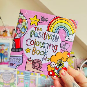 May include: A pink colouring book with a rainbow, a gumball machine, a snail, and a clown. The title is "The Positivity Colouring Book Vol. 2".