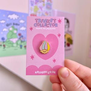 May include: A pink card with the text "TRINKET COLLECTOR" and "ENAMEL PIN COLLECTION" features a heart-shaped pin with a colorful flame design. The pin has pink, yellow, and blue colours. The card also has the text "KELLYLOU.CO.UK".