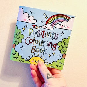 May include: A blue and green colouring book with a rainbow, clouds, and a sun. The title of the book is "The Positivity Colouring Book".