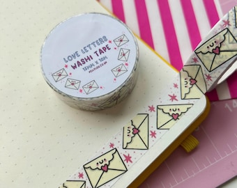 May include: A roll of yellow washi tape with a white background and pink stars. The tape features a repeating pattern of white envelopes with pink hearts and smiling faces. The tape is labeled "LOVE LETTERS WASHI TAPE 15mm x 10m #KELLYLOH.CO.UK"
