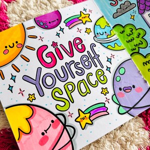 May include: A colourful illustration with the text "Give Yourself Space" surrounded by planets, stars, and rainbows. The planets have cute faces and are drawn in a cartoon style.