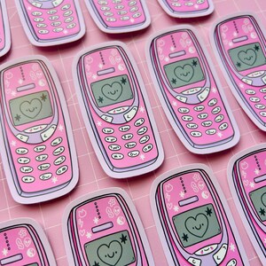 May include: Pink retro cell phone stickers with a heart-shaped smiley face on the screen. The stickers have a white border and are decorated with stars, flowers, and other small designs. The phone buttons are labeled with numbers and symbols.